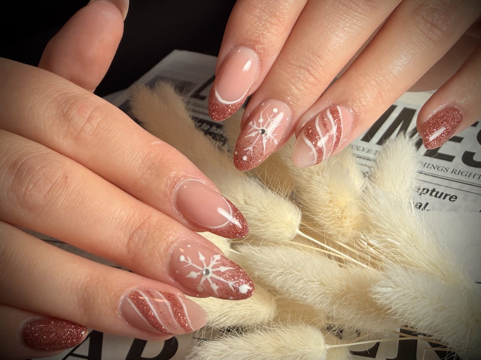 Nude-roze nagels met koperglitter en witte sneeuwvlokjes, tussen pampasgras en een krant