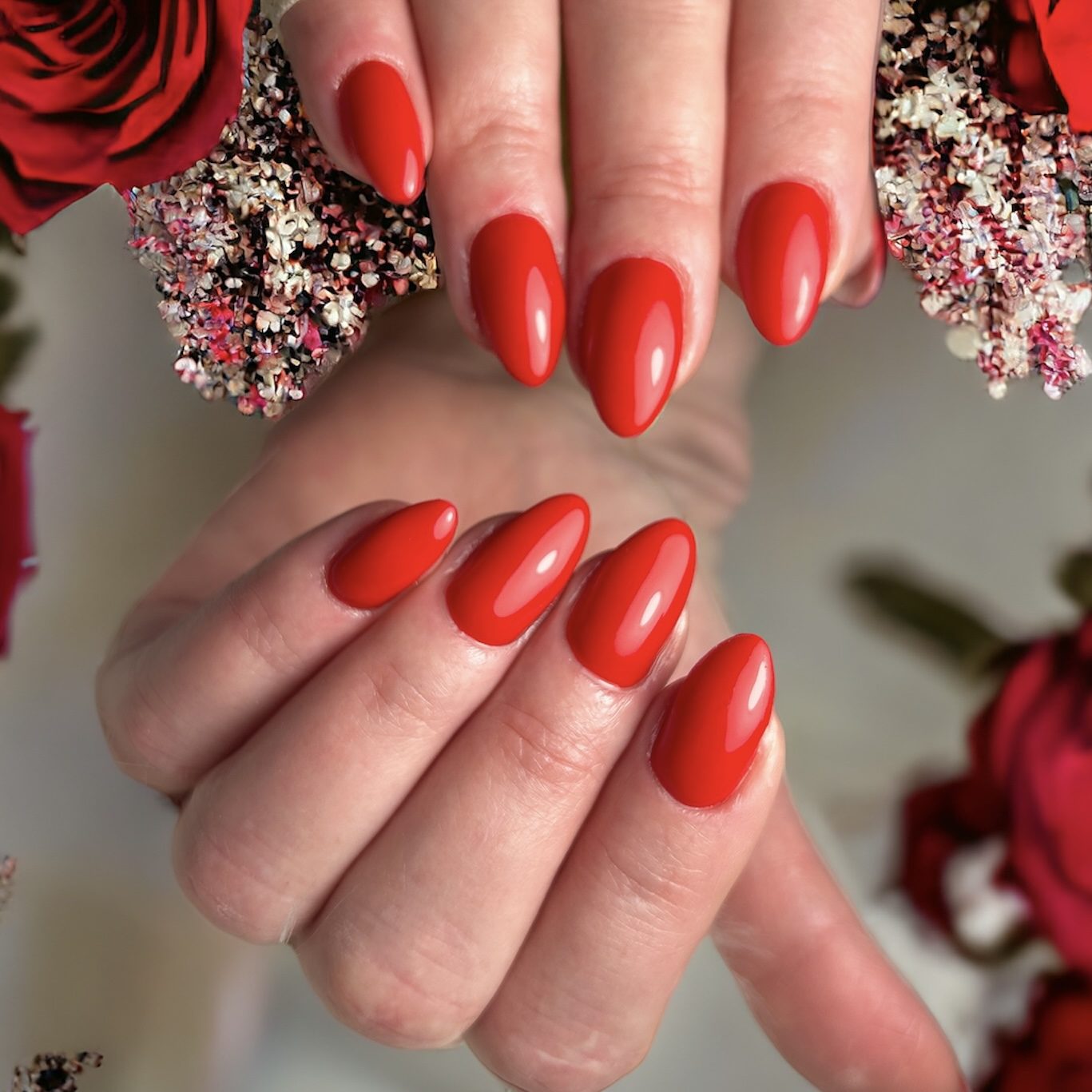 Twee handen met glanzend rode amandelnagels tussen rode rozen en droogbloemen