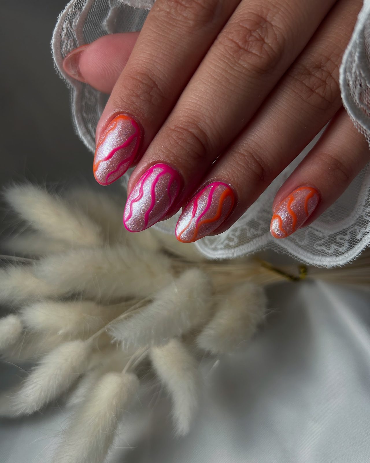 Nude amandelnagels met een marmerswirl in roze en oranje, naast kant en pampasgras