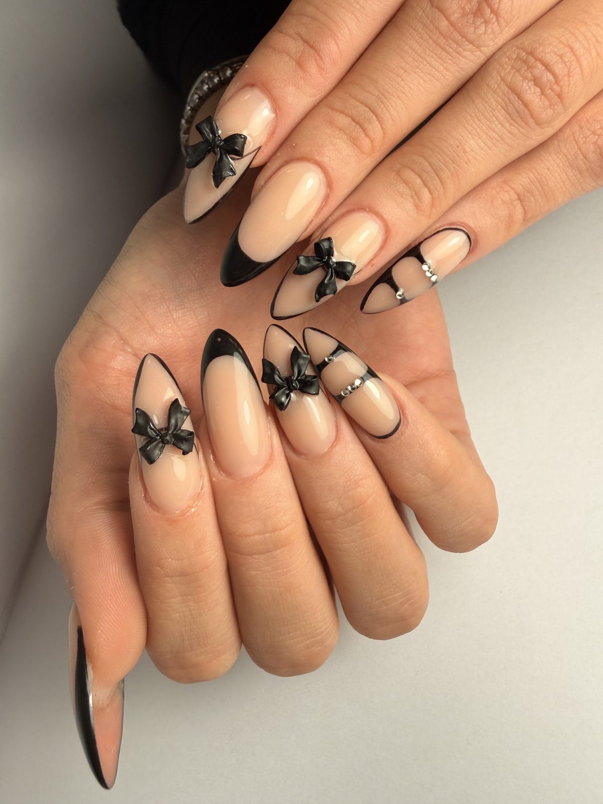 Lange nude amandelnagels met zwarte French-puntjes en zwarte strikjes in 3D