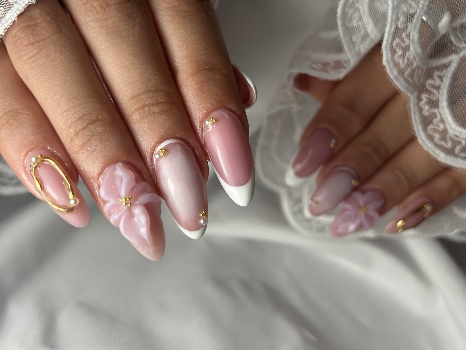 Nude amandelnagels met witte French, een gouden draadlus, parels en een roze 3D-bloem, op kant